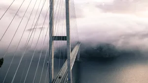 A dramatic view of a fog-covered suspension bridge with cables and the ocean below. jigsaw puzzle