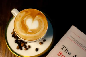 A cup of latte with heart art surrounded by coffee beans next to a book. jigsaw puzzle