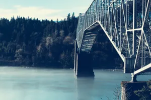 Steel suspension bridge crossing a fog-covered river with forest background. jigsaw puzzle